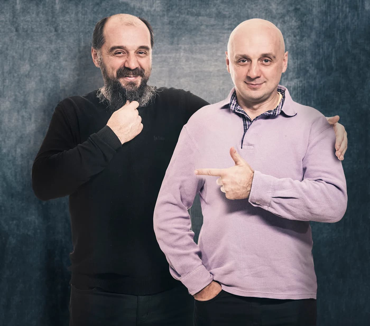 Rotaļīgs korporatīvs portrets diviem vīriešu kolēģiem, smaidot un rādot vienam uz otru, uz pelēka fona