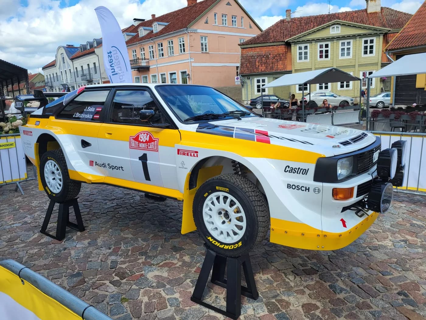 Oriģinālais foto dzeltenam un baltam Audi rallija auto uz bruģa laukuma ar ēkām fonā